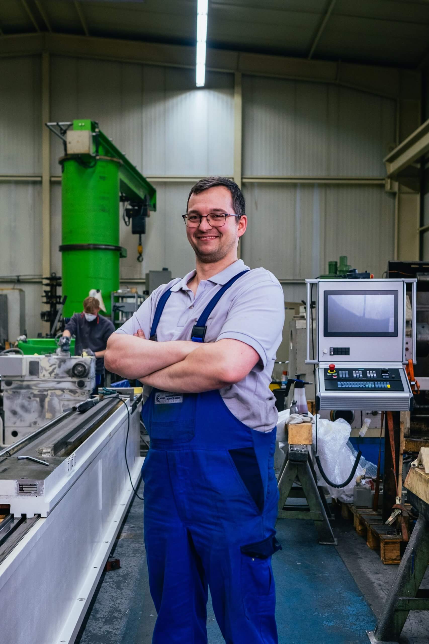 Ein Mitarbeiter steht mit verschränkten Armen in der Werkstatt vor Maschinen und einem Steuerpult, bei einem Maschinenbau Unternehmen Aschersleben.