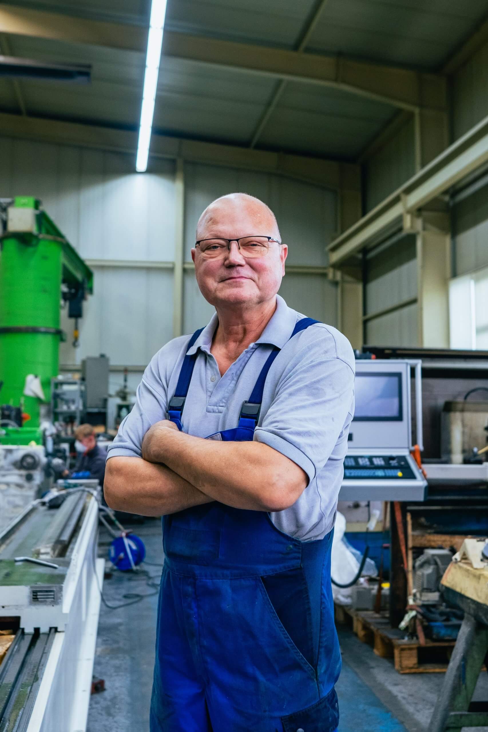 Der Eigentümer steht in Arbeitskleidung mit verschränkten Armen in der Werkstatt, Maschinen und Steuerpult im Hintergrund, bei einem Maschinenbau Unternehmen in Aschersleben.